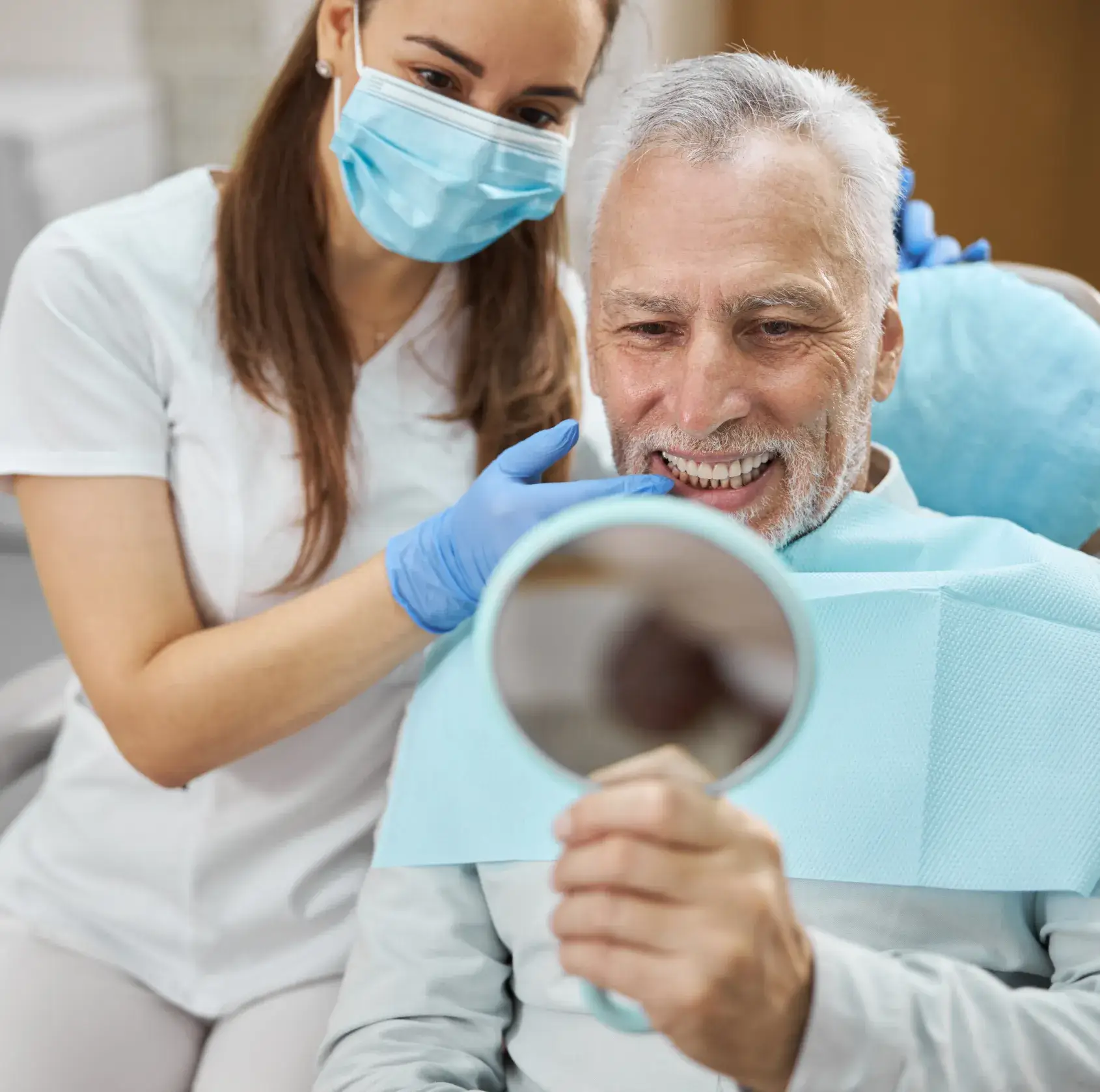 Senior man smiling and checking his teeth in a handheld mirror with a dentist beside him.