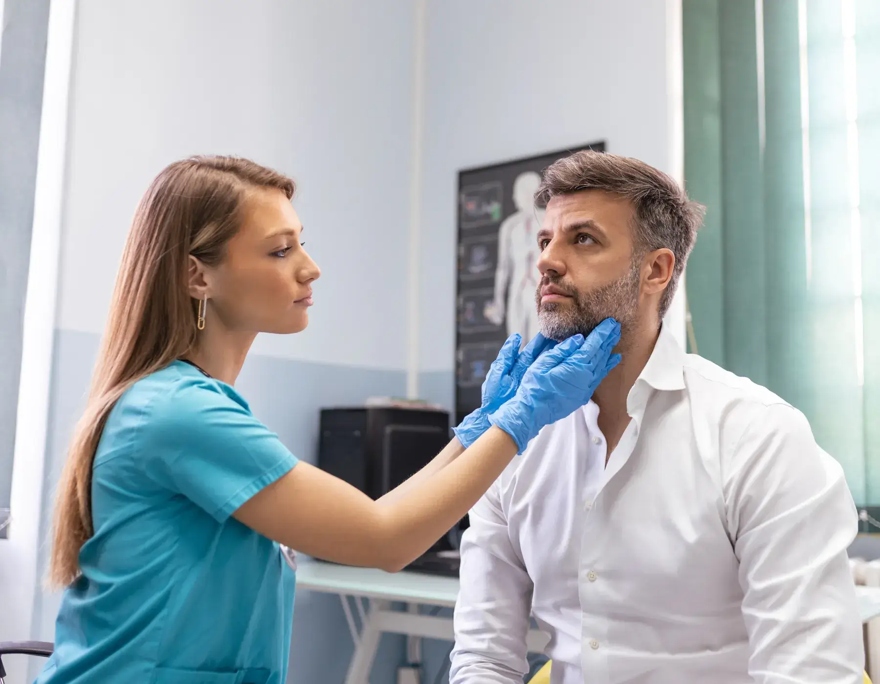 Woman healthcare professional in blue scrubs examining a man's neck in a medical office.