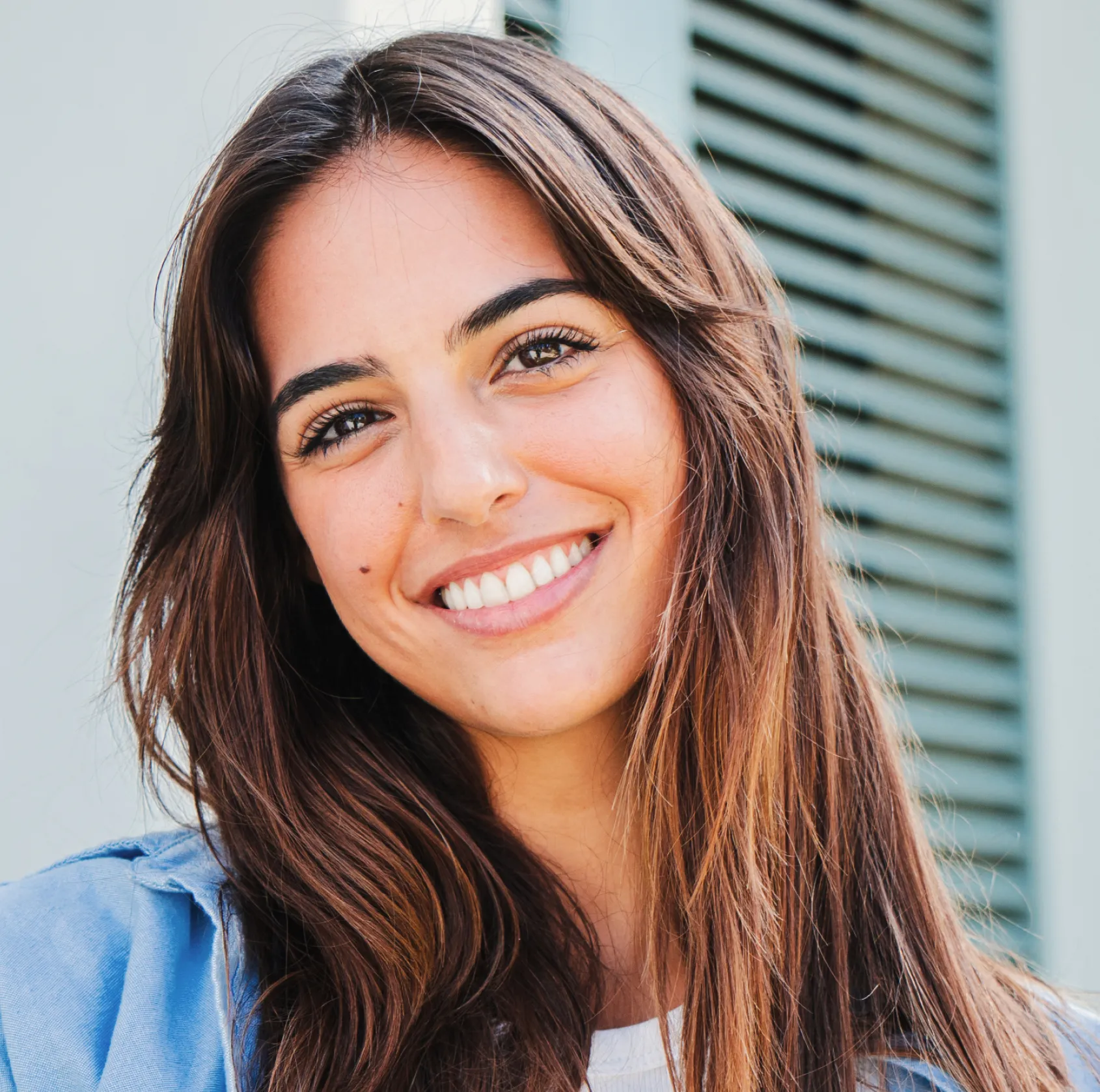 Close-up of a smiling woman with long brown hair and clear skin wearing a light blue top.