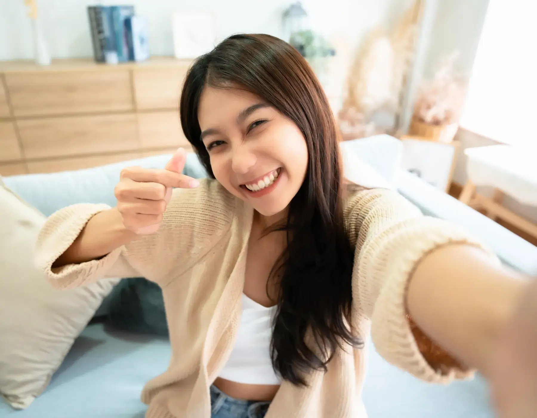 Smiling woman taking a selfie indoors, making a finger heart gesture with her hand.