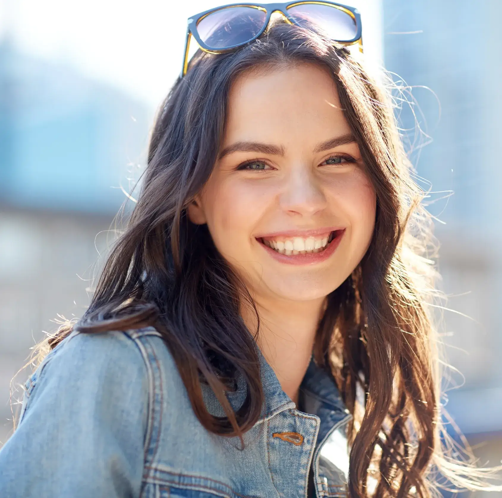 Young woman with long dark hair wearing sunglasses on her head and a denim jacket smiling outdoors.