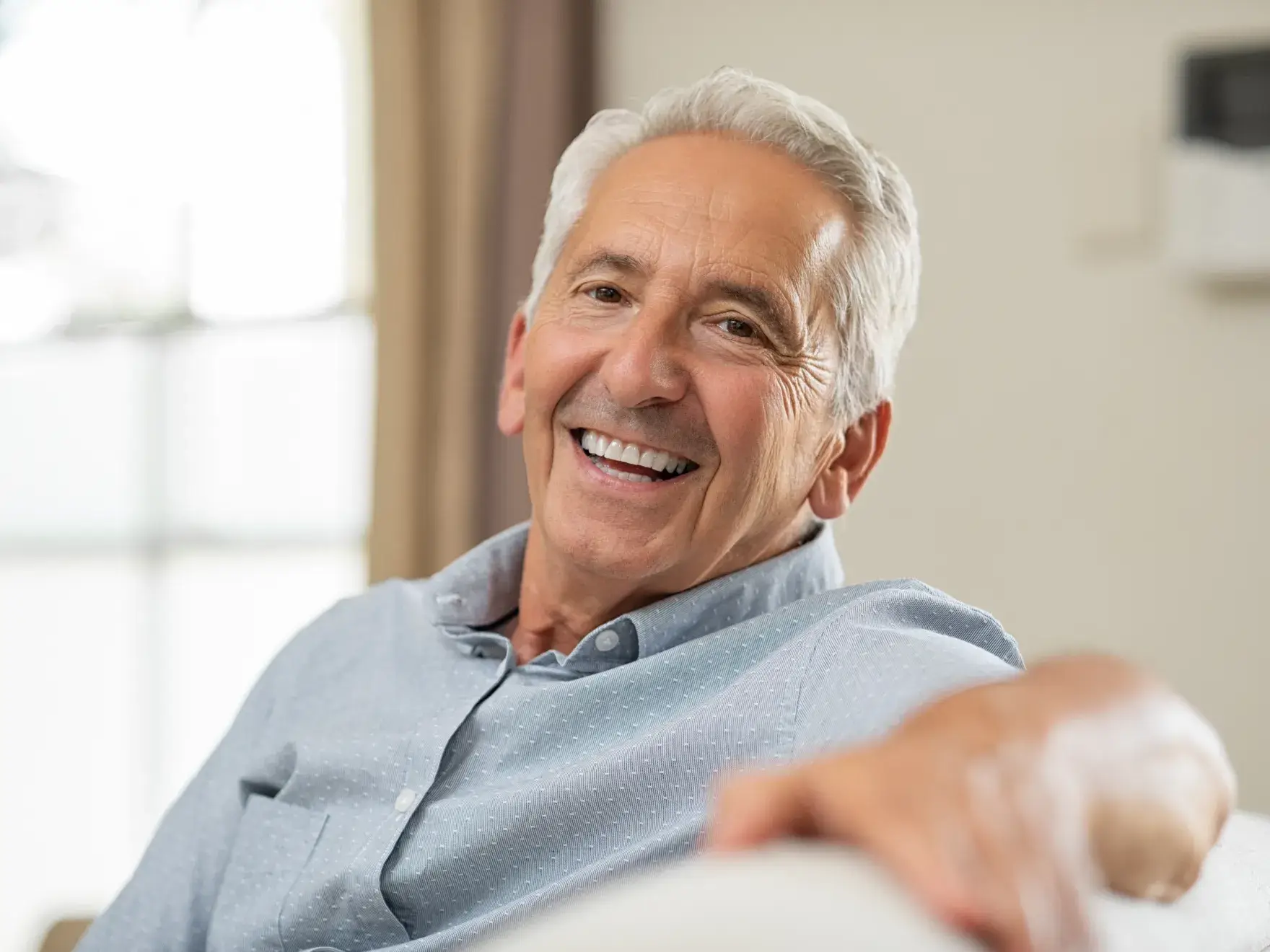 Smiling older man with gray hair wearing a light blue shirt sitting indoors on a couch.