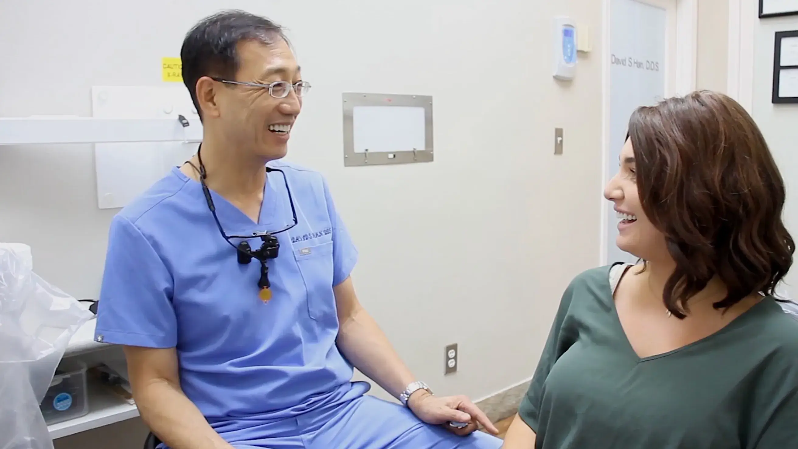 Male dentist in blue scrubs smiling and talking to a female patient in a dental office.
