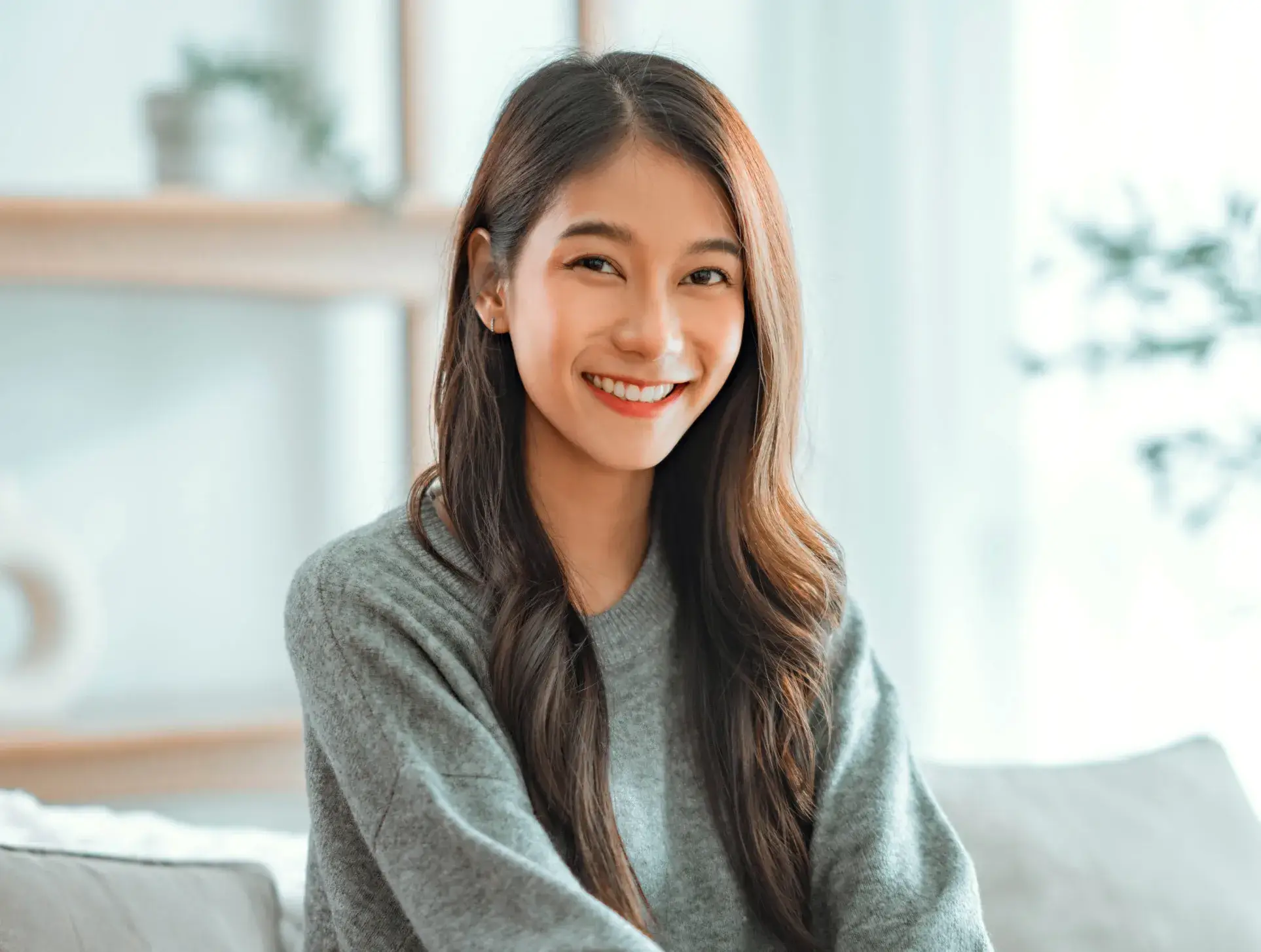 Smiling young woman with long hair wearing a gray sweater in a bright, cozy room.